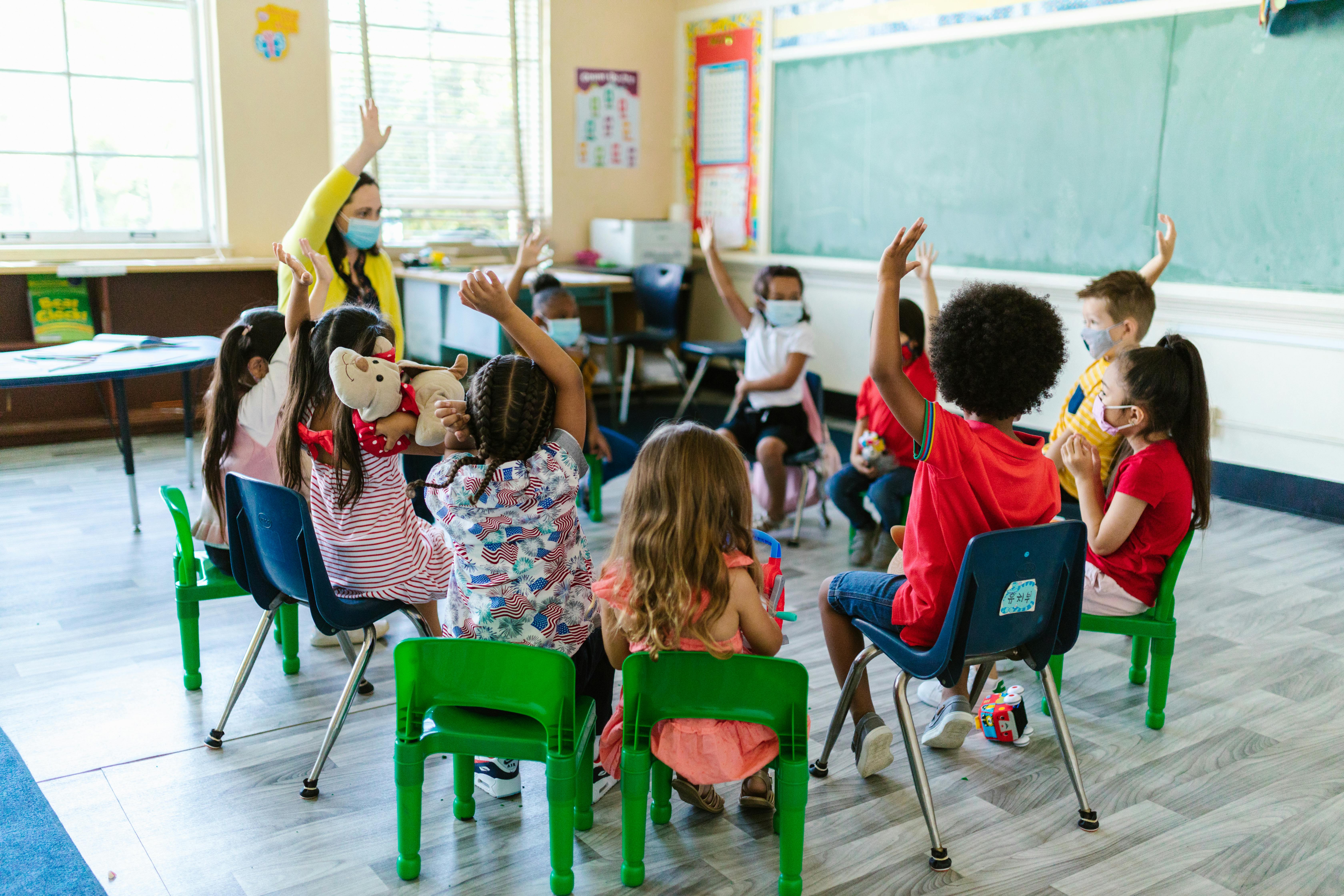 Masked young children sitting in a classroom circle raising their hands with a masked teacher also raising her hand.