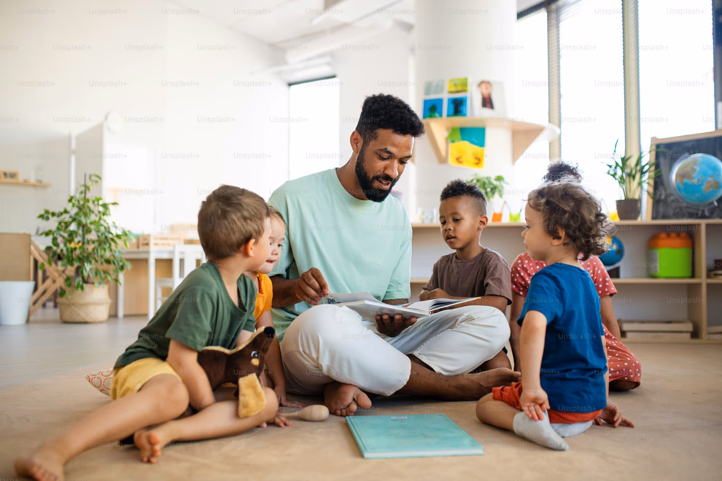 Male teacher sitting cross-legged on the floor reading a book to a group of young children in a bright classroom.