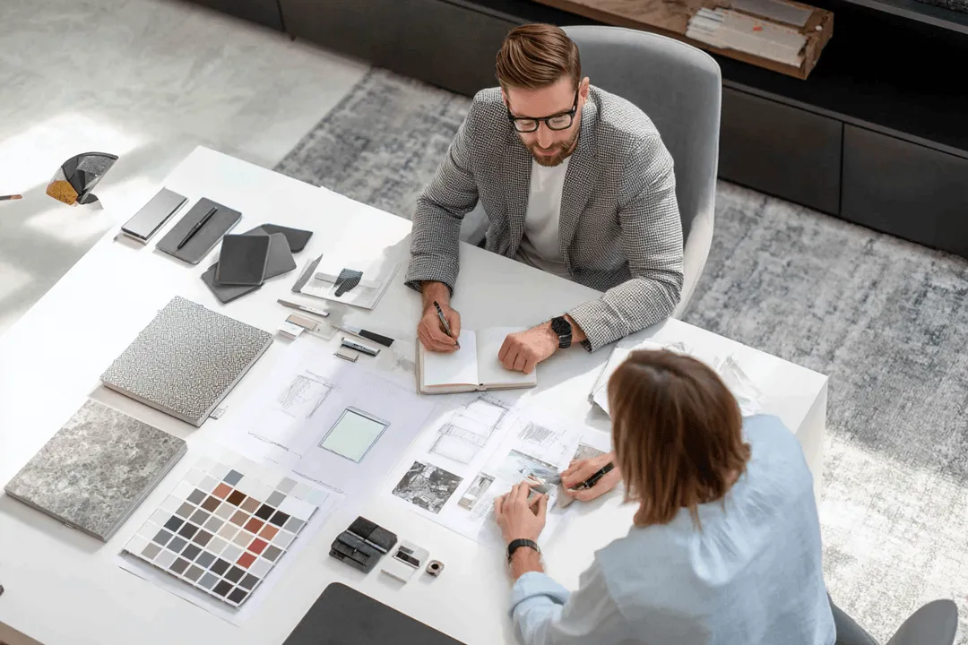 A clean and modern interior design consultation scene focused on custom mirror design. A professional consultant and a client sit at a design table reviewing mirror samples and sketches.