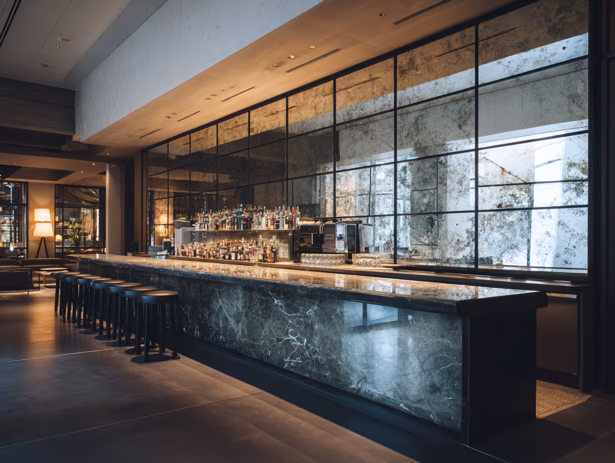 Modern bar interior with a long marble counter, black stools, backlit shelves of liquor bottles, and large antique-style mirrors.