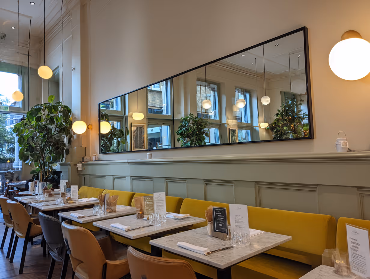 Modern restaurant interior with marble tables, yellow banquette seating, brown chairs, large rectangular mirror, and hanging globe lights.