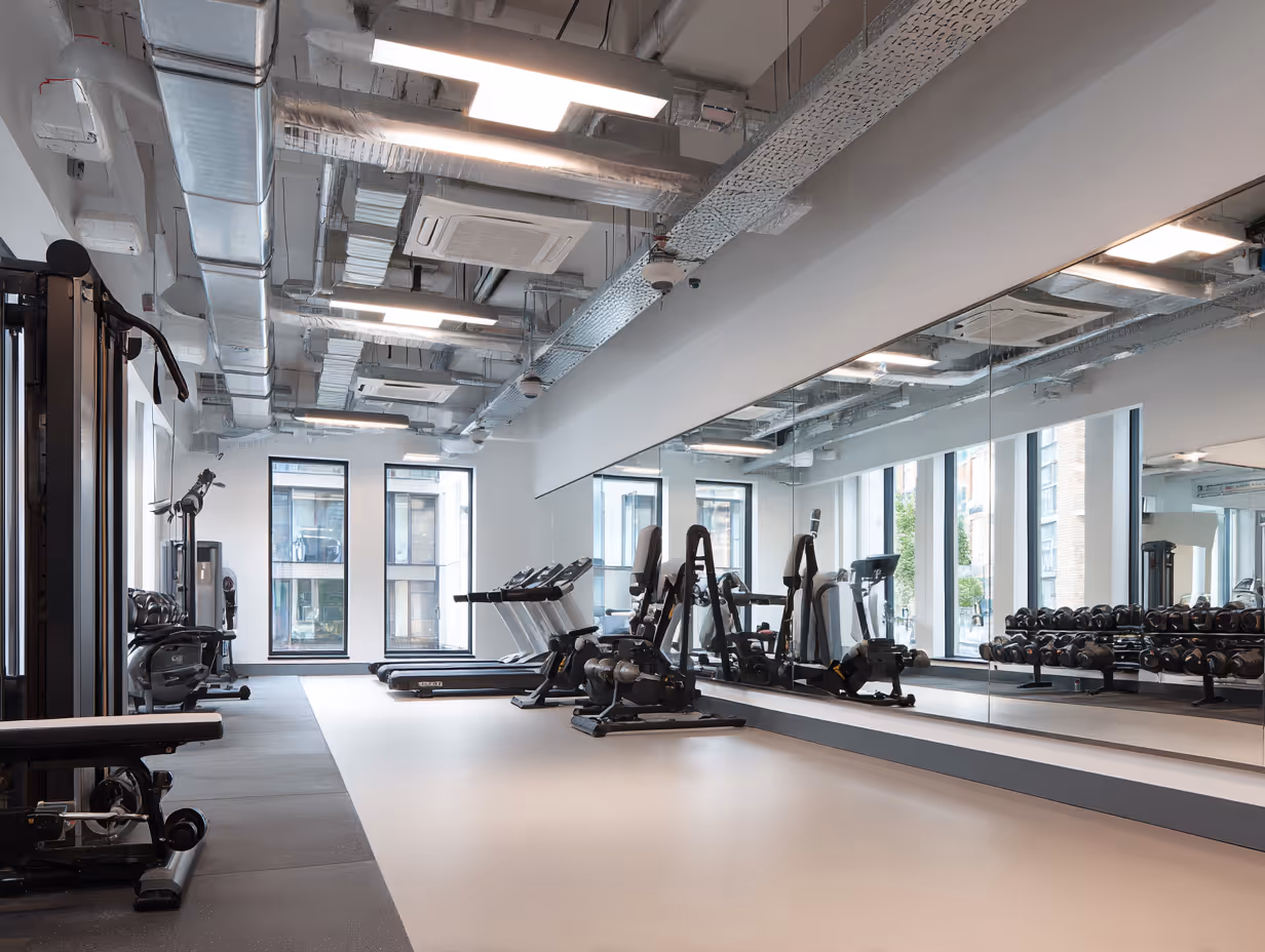 Modern gym with treadmills, weight machines, dumbbells, large mirrors, and industrial ceiling piping.