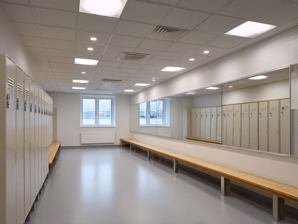 Modern school locker room with beige lockers along the left wall and a long wooden bench beneath a large mirror on the right wall.