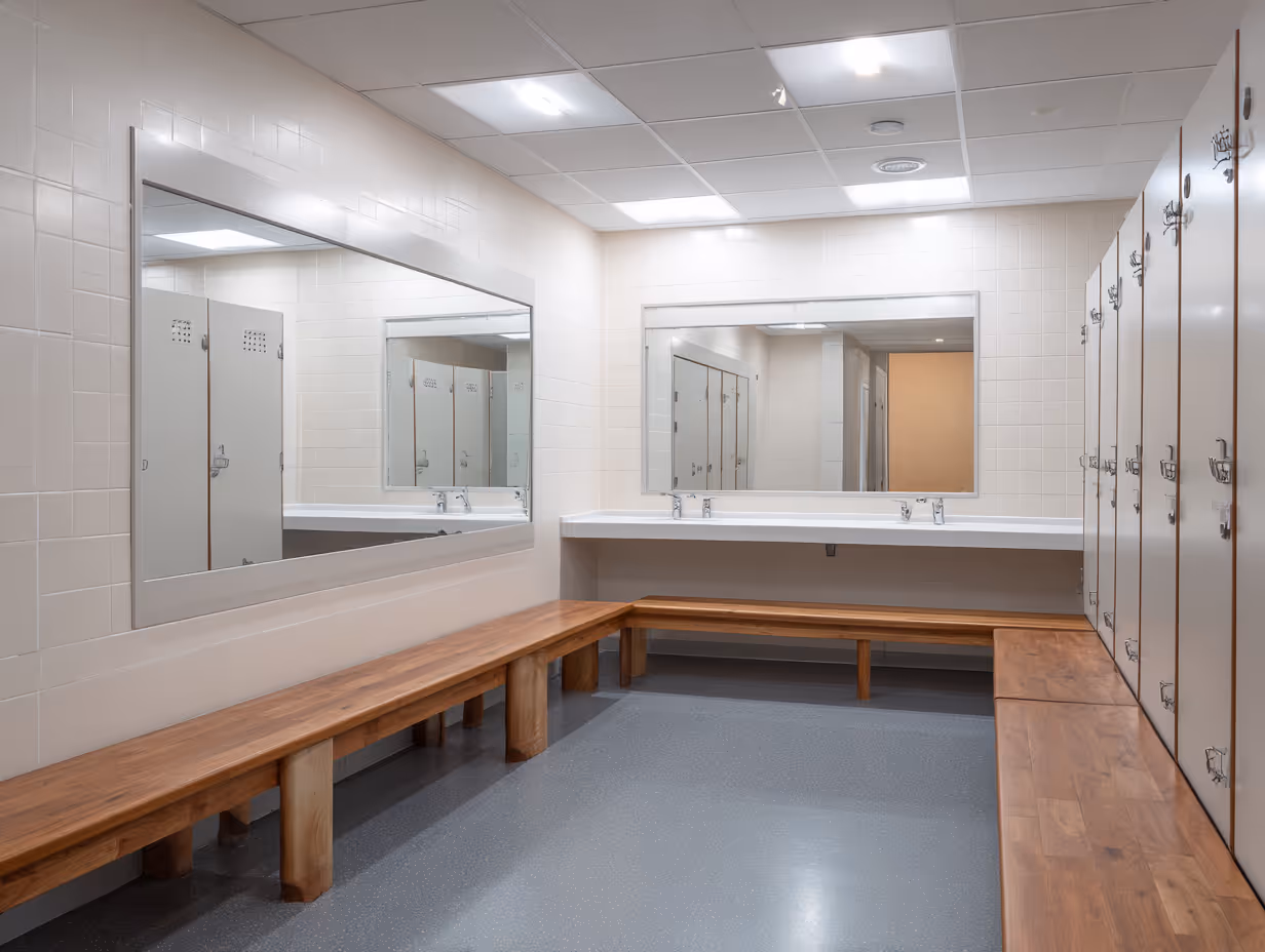 Empty locker room with wooden benches, white lockers, two large mirrors, and sinks beneath the mirrors.