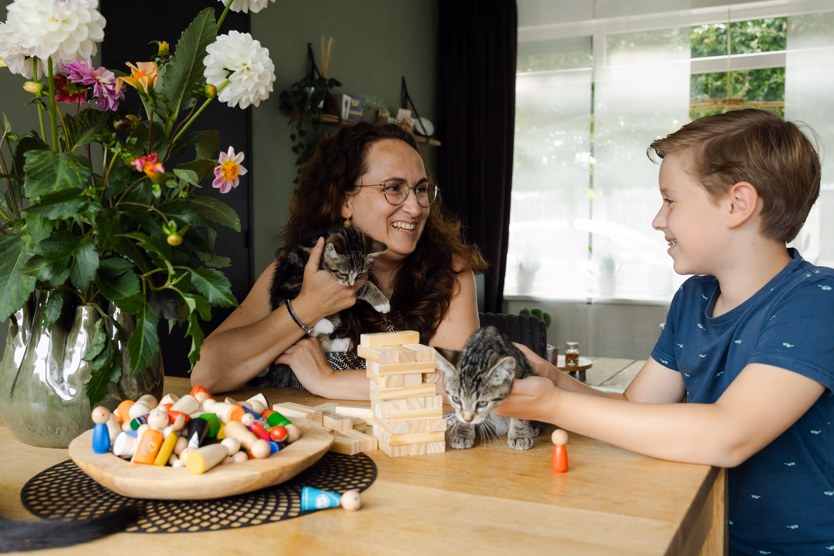 Een vrouw en een jongen die lachend naar elkaar kijken terwijl ze kittens vasthouden, naast een gestapeld houten blokkespel op een tafel.