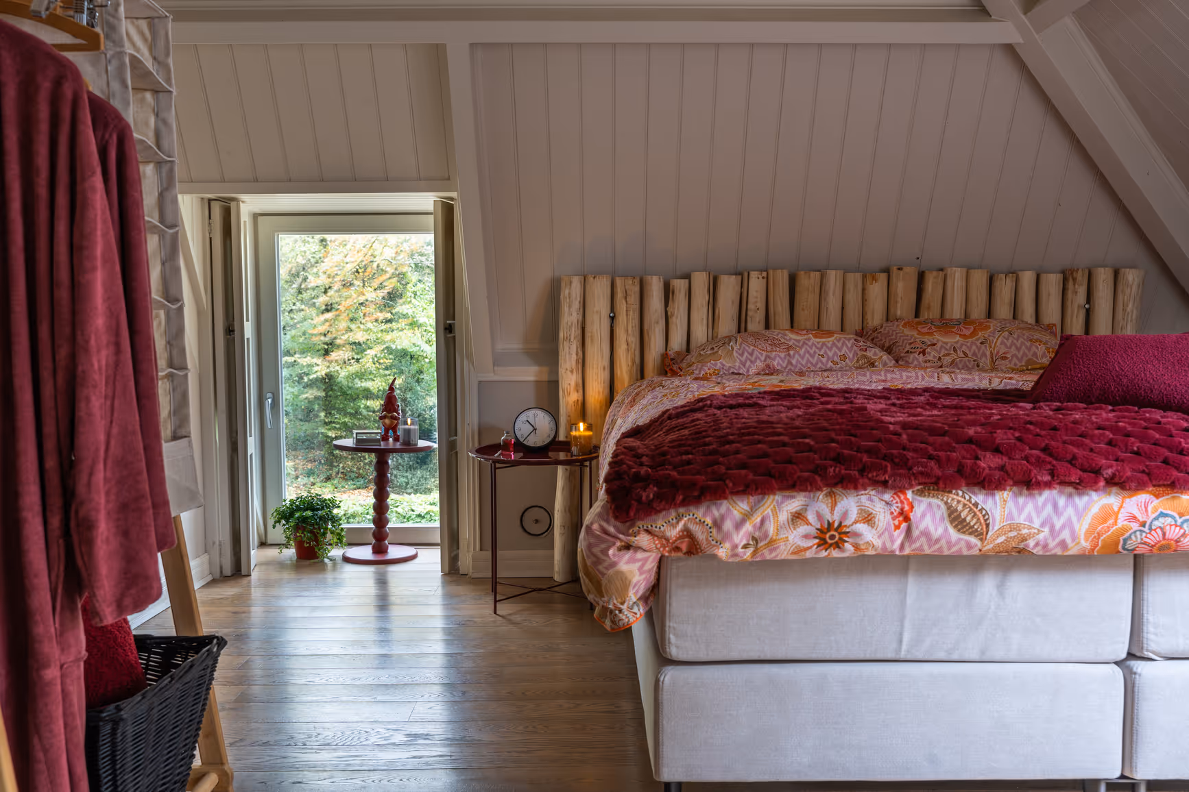 Slaapkamer met een gebloemd bed, houten hoofdbord, nachtkastje met klok en kaarsen, en een deur met uitzicht op groen.