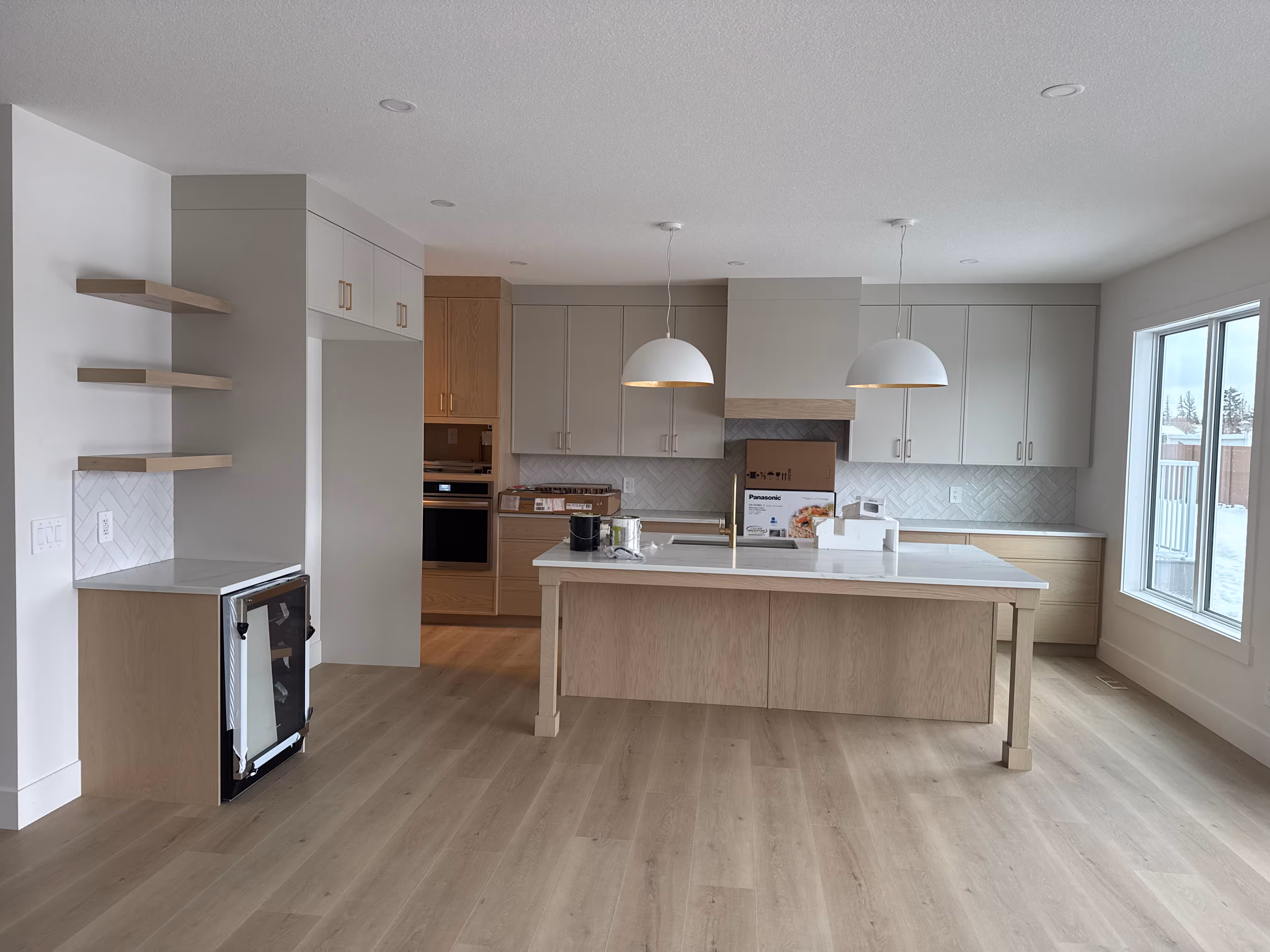 Modern kitchen with light wood flooring, a central island with white countertop, two pendant lights, and large window overlooking a snowy yard.