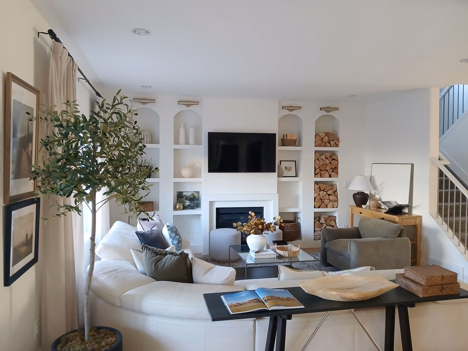 Bright, modern living room with white curved sofa, gray armchair, glass coffee table, wall-mounted TV, built-in shelves with decor, and indoor potted tree.