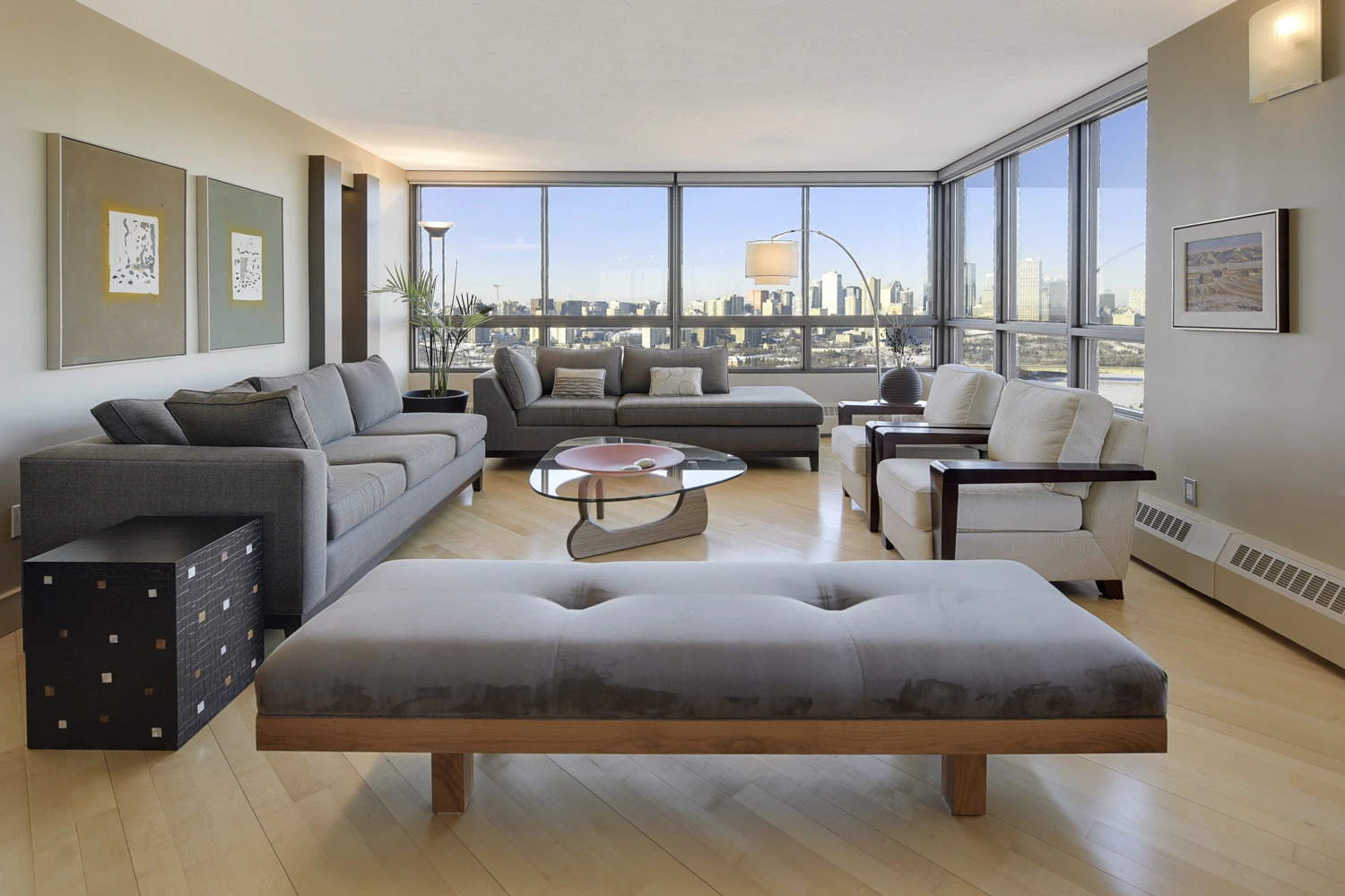 Modern living room with large windows showing city skyline, featuring gray sofa, chaise lounge, white armchairs, and glass coffee table.