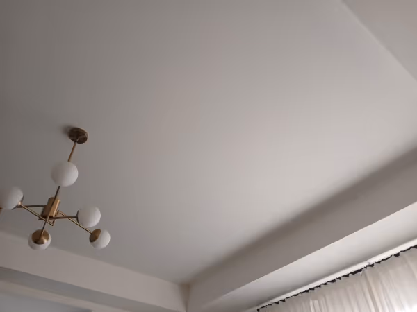 White ceiling with a modern brass chandelier featuring six round white bulbs and beige curtains partially visible at the bottom right.