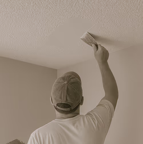 Man wearing a cap applying a patch to a textured ceiling using a putty knife.