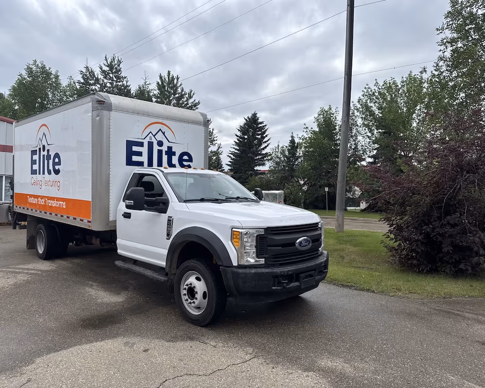 White Ford box truck with Elite Ceiling Texturing logo parked on a road near trees and a utility pole under a cloudy sky.