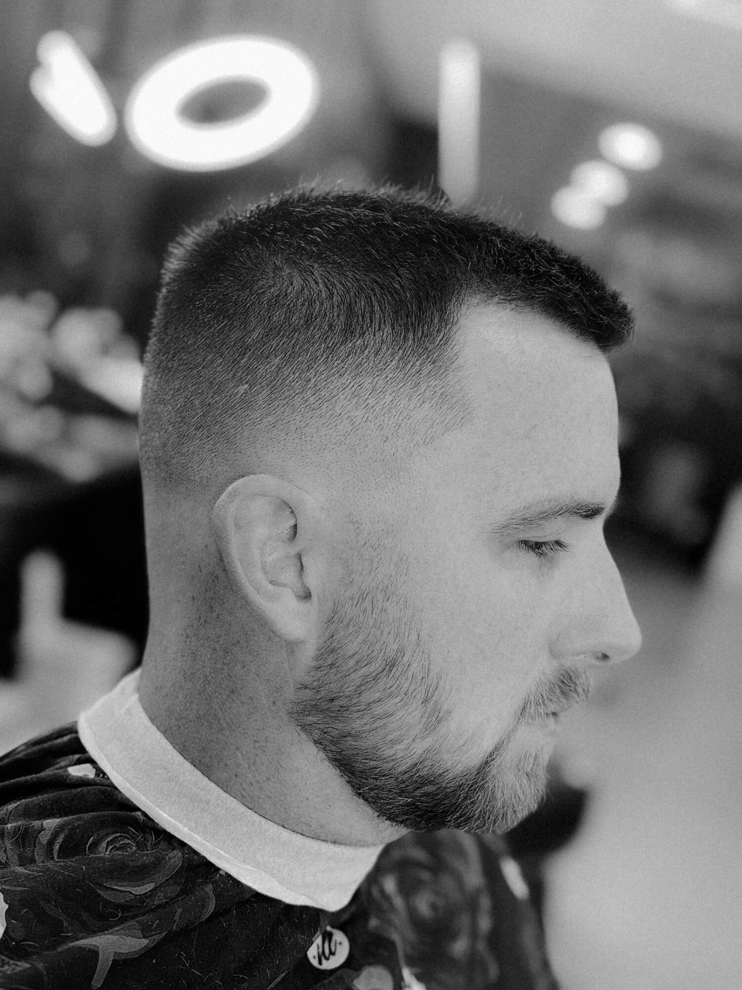 Side profile of a man with a short fade haircut and neatly trimmed beard in a barbershop.