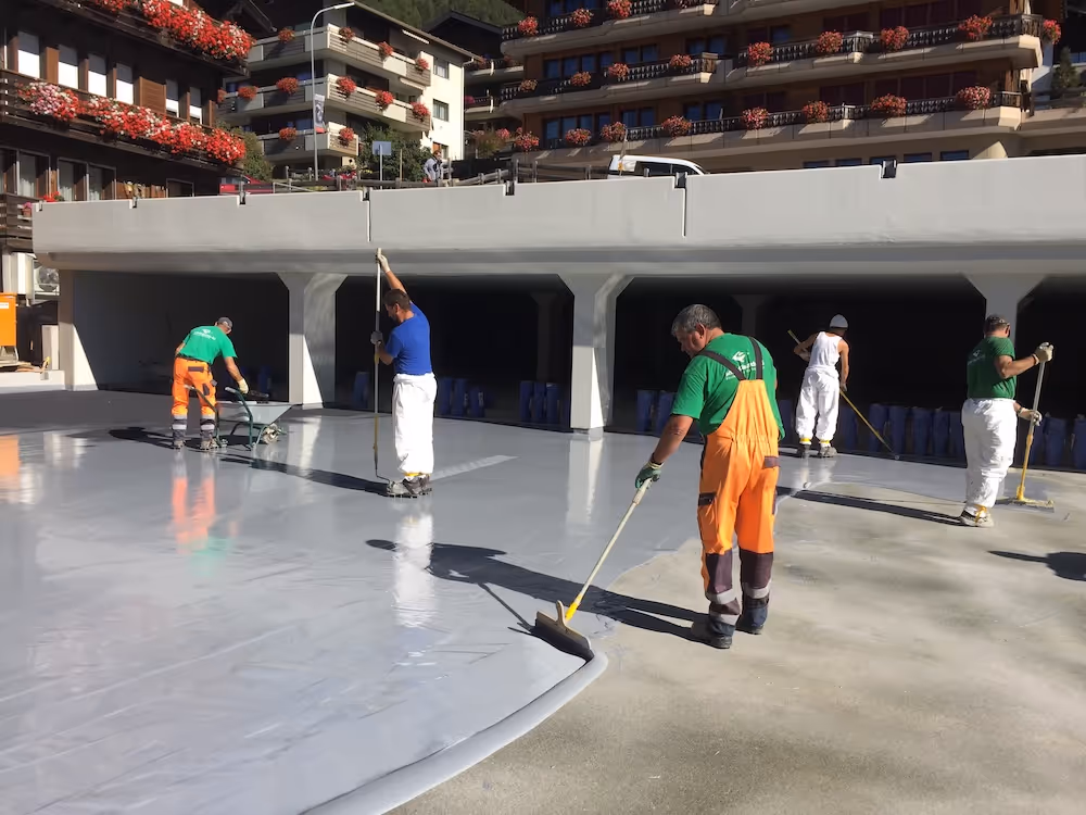 Fünf Arbeiter in Arbeitskleidung tragen eine graue Beschichtung auf einem großen Außenboden vor einem Gebäude mit Balkonen und roten Blumen auf.
