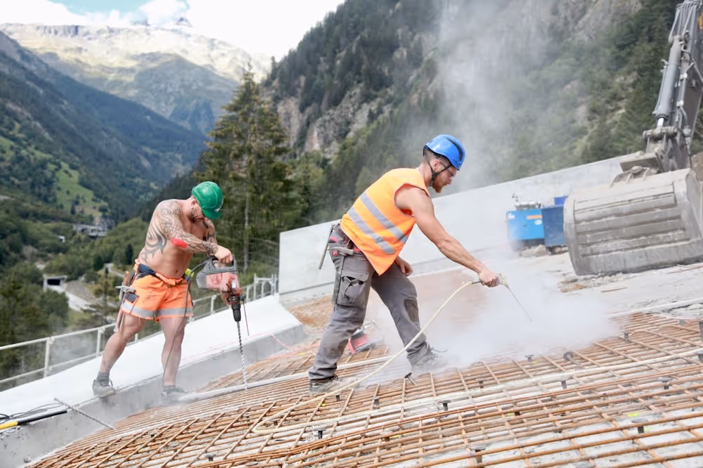 Zwei Bauarbeiter mit Schutzhelmen bearbeiten eine große Stahlarmierung auf einer Baustelle in den Bergen.