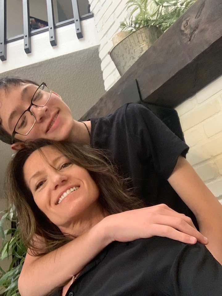 Smiling woman with brown hair hugged from behind by a boy wearing glasses and a black shirt indoors.