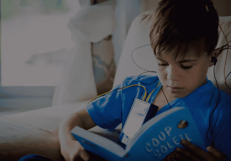 Young boy wearing earphones reading a blue book titled 'Coup Soleil' while sitting on a couch.