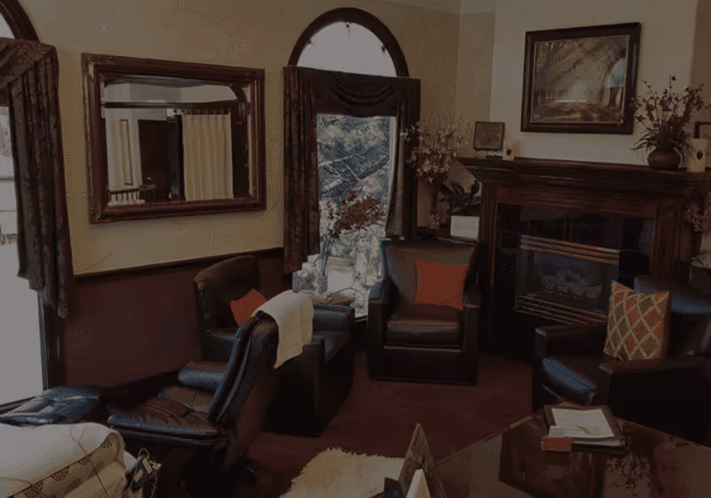 Cozy living room with dark leather chairs, a fireplace, decorative pillows, and floral arrangements near a large window with curtains.