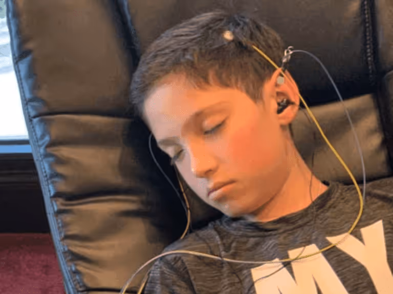 Boy sitting in a chair with closed eyes wearing hearing test earphones connected by wires.