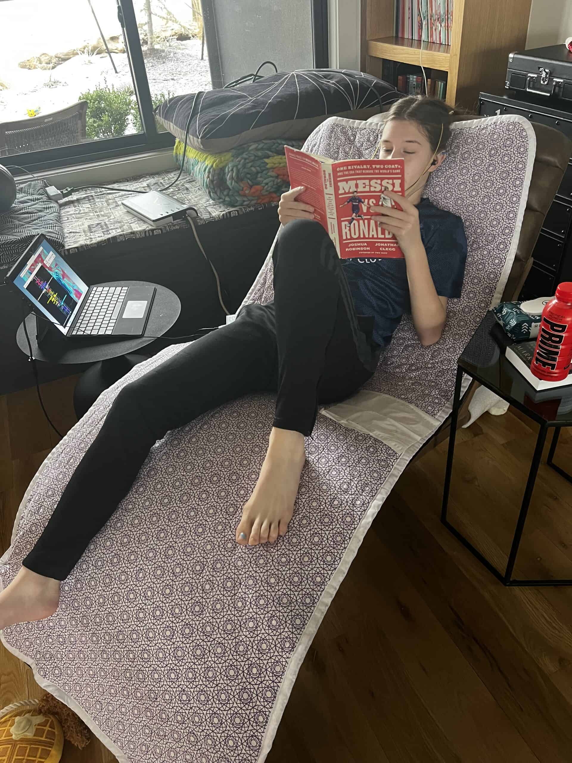 Young person reclining on a patterned lounge chair reading a book titled 'Messi vs Ronaldo' with a laptop on a nearby table and a red water bottle.