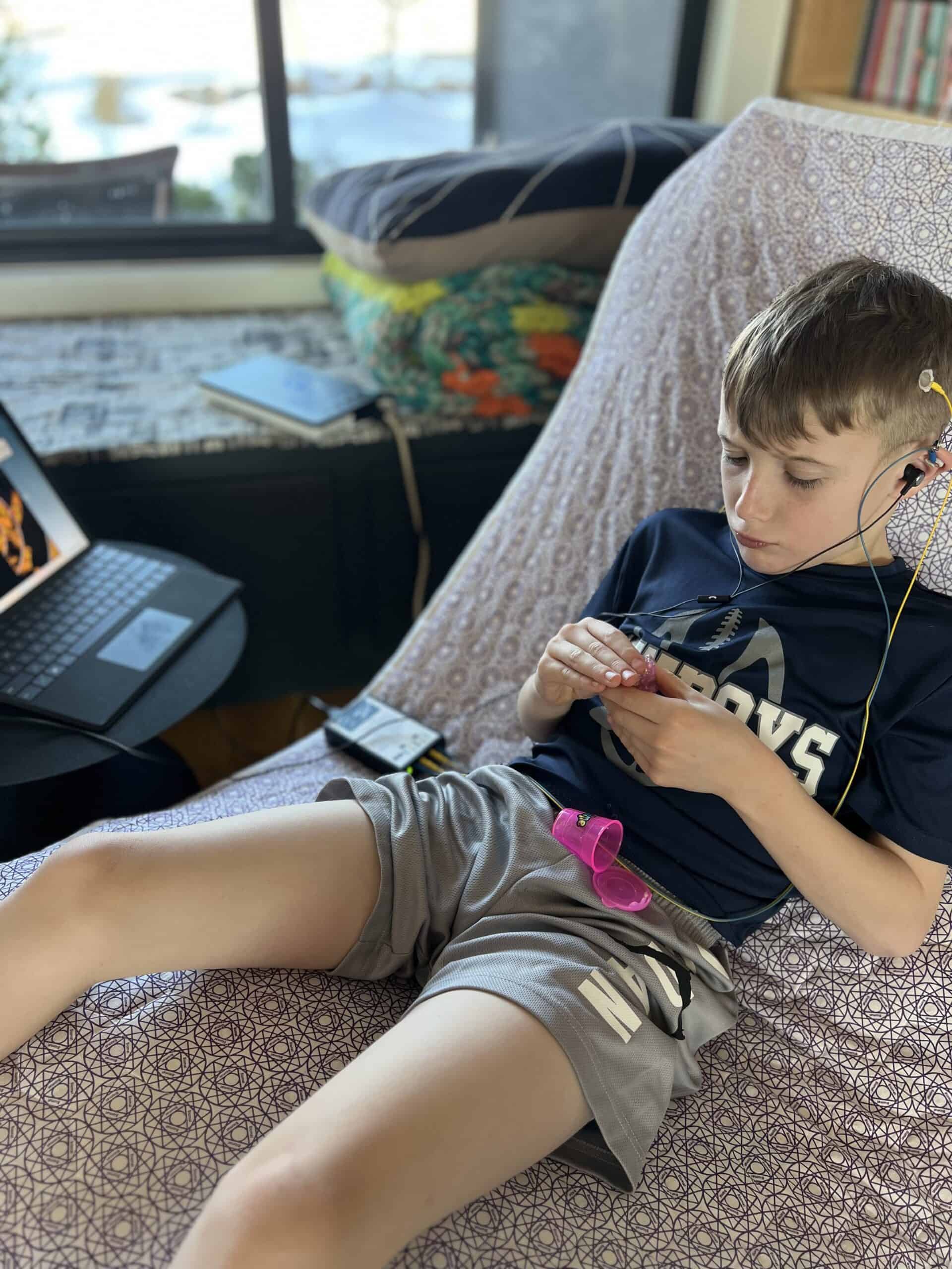 Boy lying on a patterned blanket, wearing earphones and a Cowboys shirt, focused on a small object in his hands.