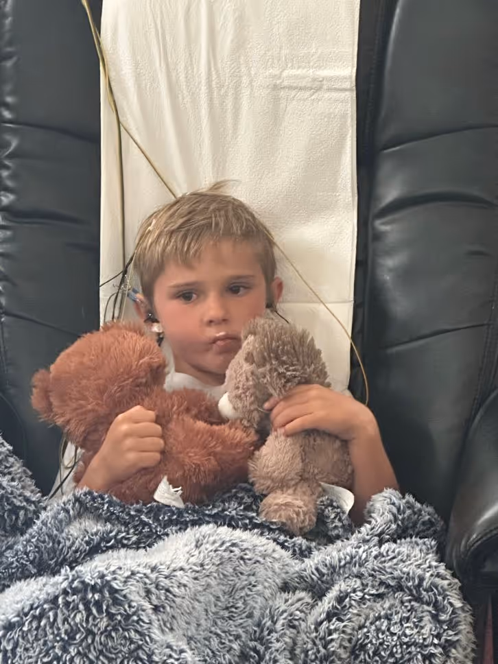 Young boy sleeping in a chair wearing a gray shirt with ear electrodes attached by wires for monitoring.