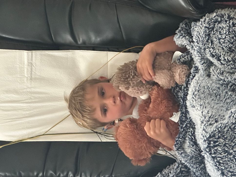 Young boy sleeping in a chair wearing a gray shirt with ear electrodes attached by wires for monitoring.