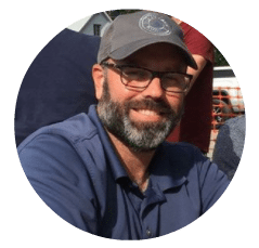 Man with glasses, beard, and mustache wearing a gray cap and blue shirt smiling outdoors.
