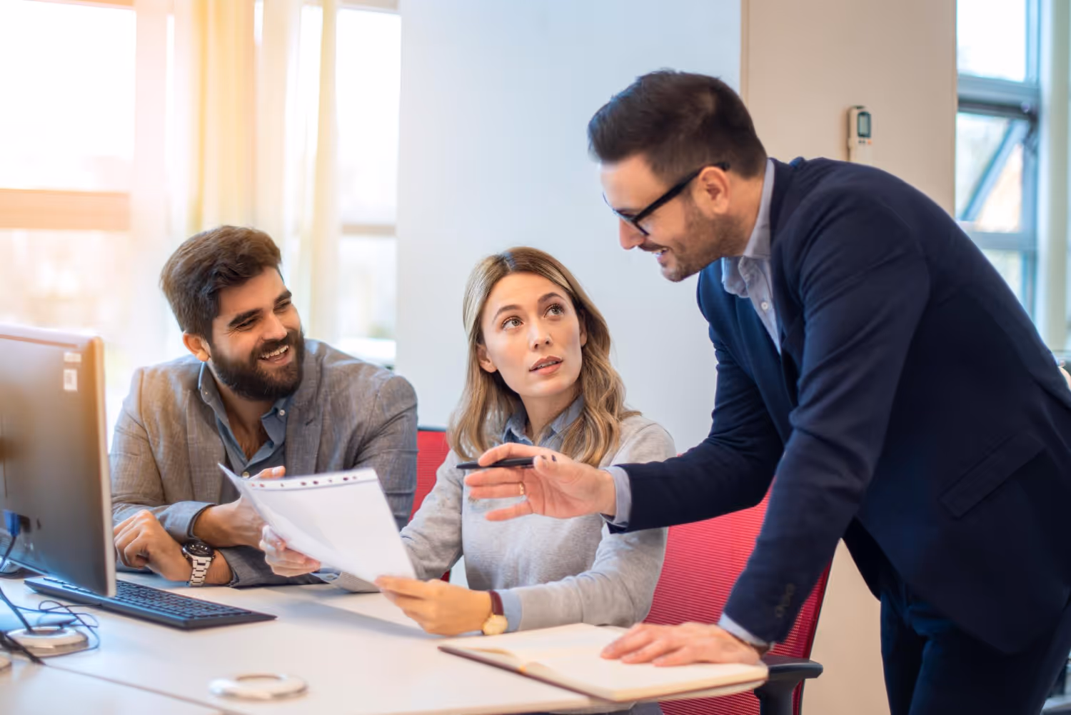 Three coworkers engaged in a discussion around a desk, one man standing and pointing with a pen, a woman holding papers, and another man smiling.