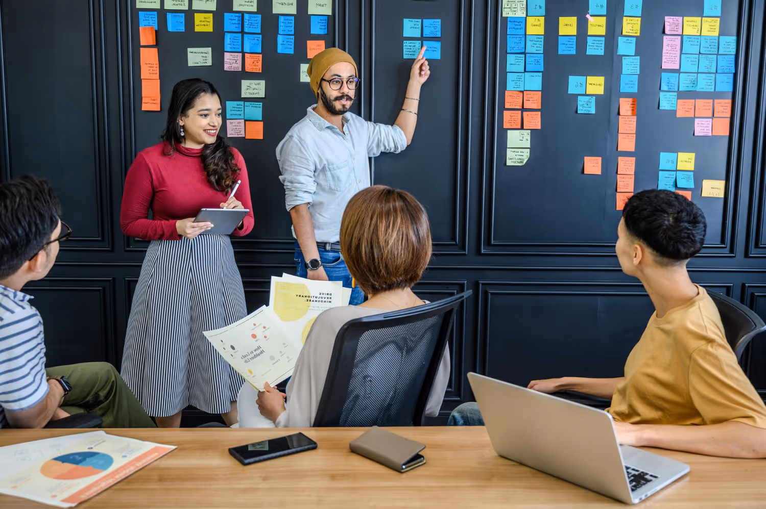 A diverse team in a meeting room with sticky notes on the black wall, one person pointing and others listening and taking notes.