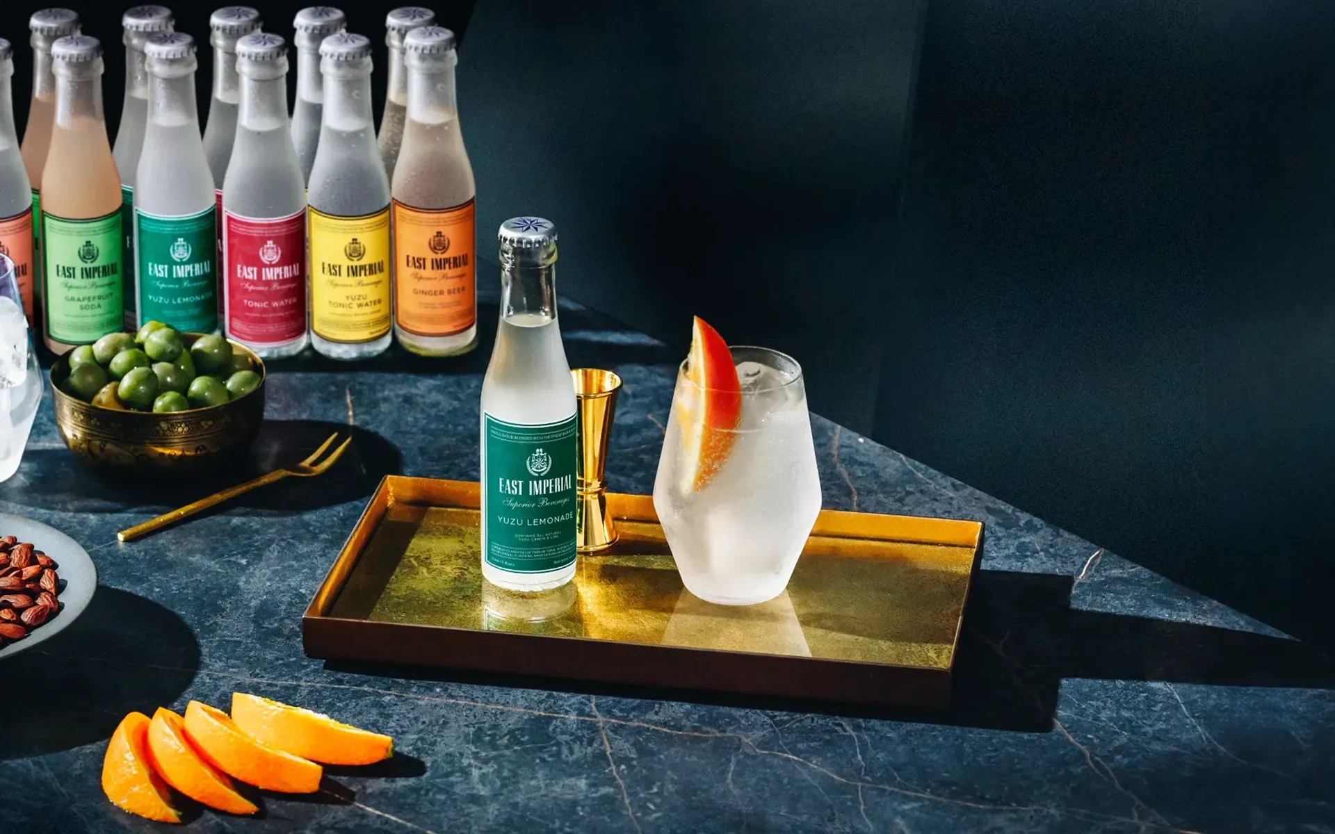 Glass of iced drink garnished with an orange slice, a bottle of East Imperial Yuzu Lemonade, and sliced oranges on a dark marble surface with additional bottles and a bowl of green olives in the background.