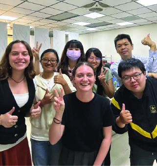 Group of seven diverse young adults smiling and giving thumbs up in an indoor setting.