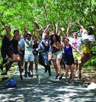 Group of diverse young adults jumping cheerfully on a tree-lined path outdoors.