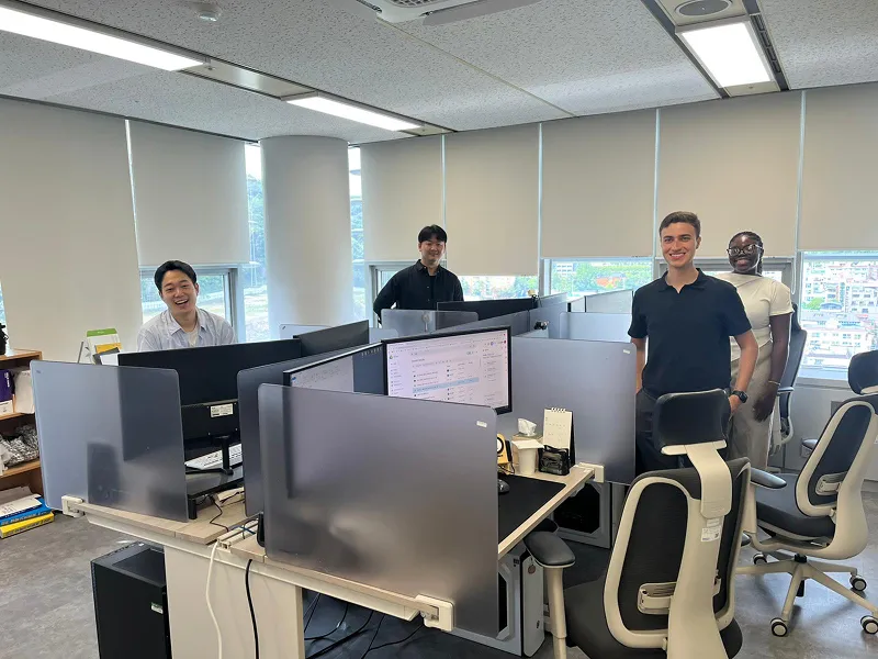 Group of seven diverse young adults smiling and giving thumbs up in an indoor setting.