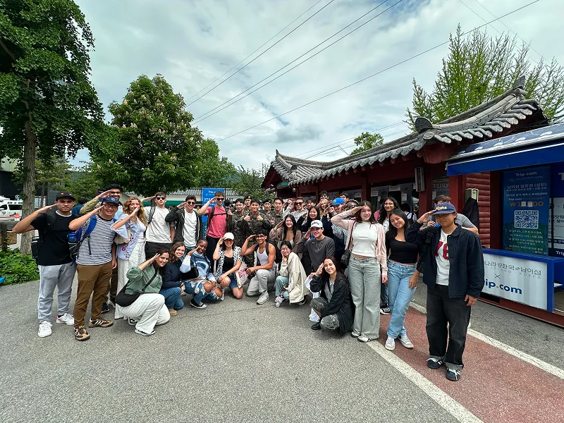Large group of diverse young people posing and smiling outdoors near traditional-style buildings and greenery.