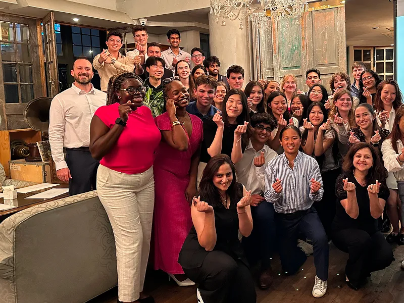 Group of six diverse young women standing together outside a building, five wearing matching navy blue t-shirts with text.