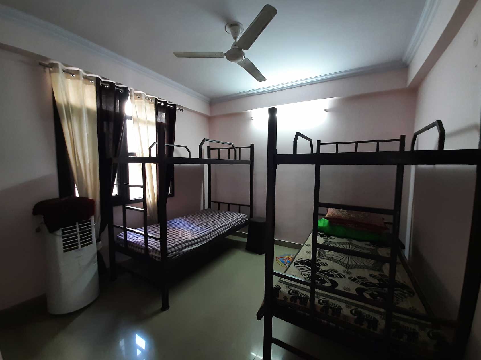 A shared dormitory with bunk beds, light-colored walls, and a ceiling fan. The room has large windows with curtains.