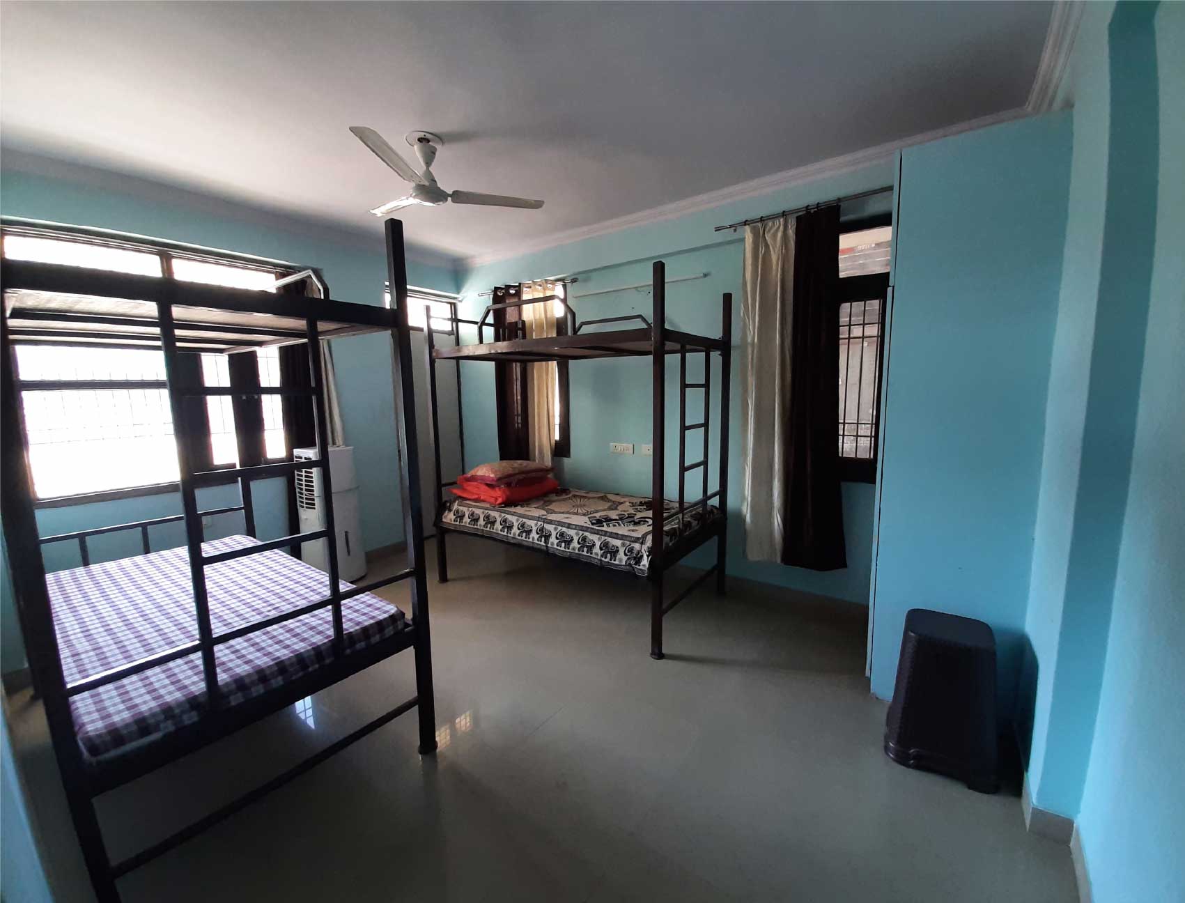 A shared dormitory room with multiple bunk beds, blue walls, and large windows for natural light.