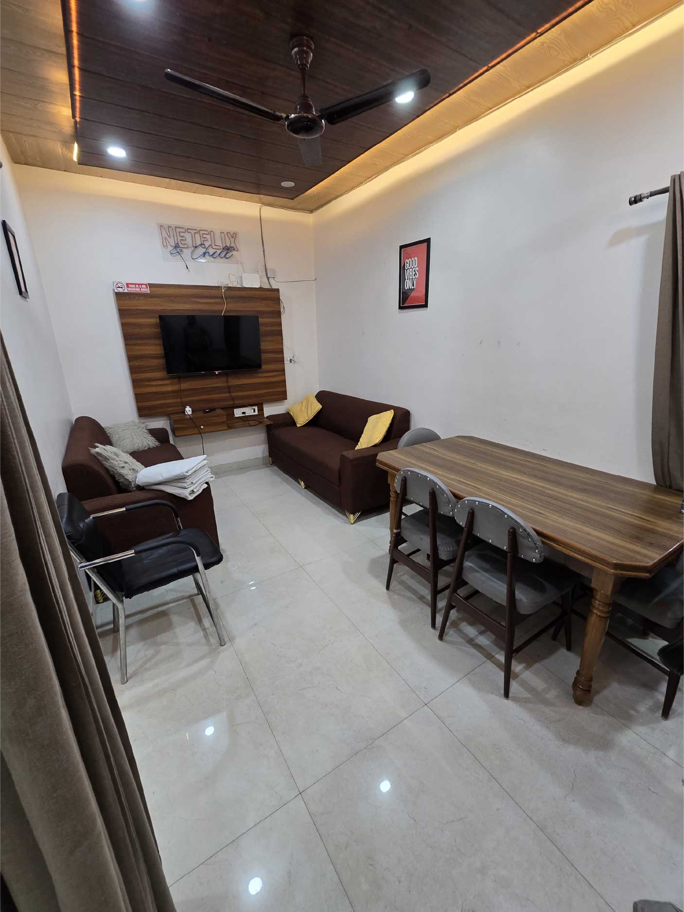 A living area with sofas, a dining table, and a mounted TV on a wooden wall panel. The ceiling has recessed lighting, and the room has a polished floor.