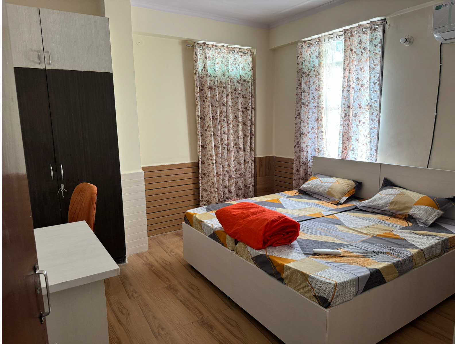 A comfortable bedroom with a double bed, colorful bedding, wooden flooring, and curtains covering large windows.