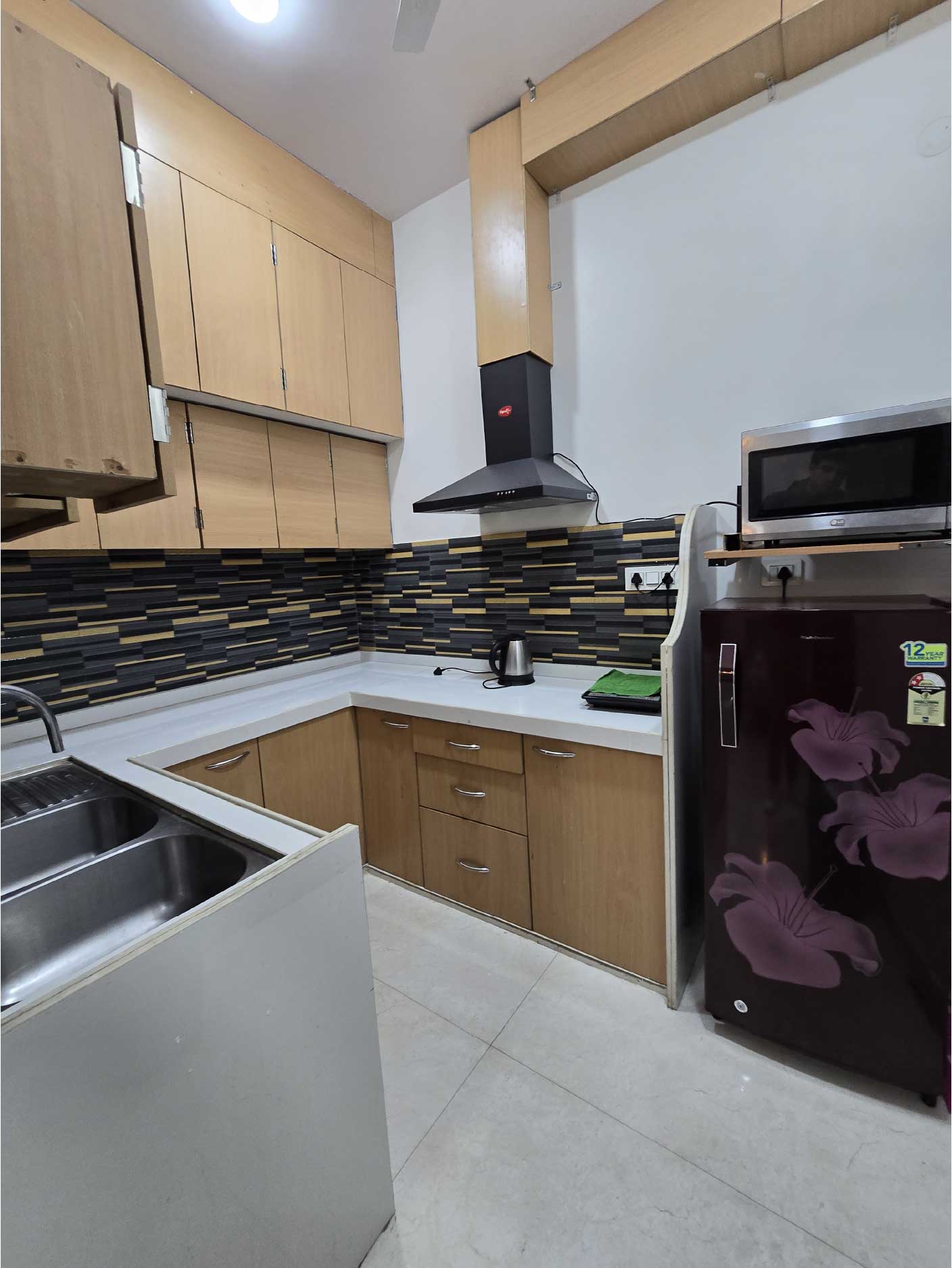 A kitchen with wooden cabinets, white countertops, and a tiled backsplash featuring black and gold accents. Appliances like a refrigerator and chimney are visible.