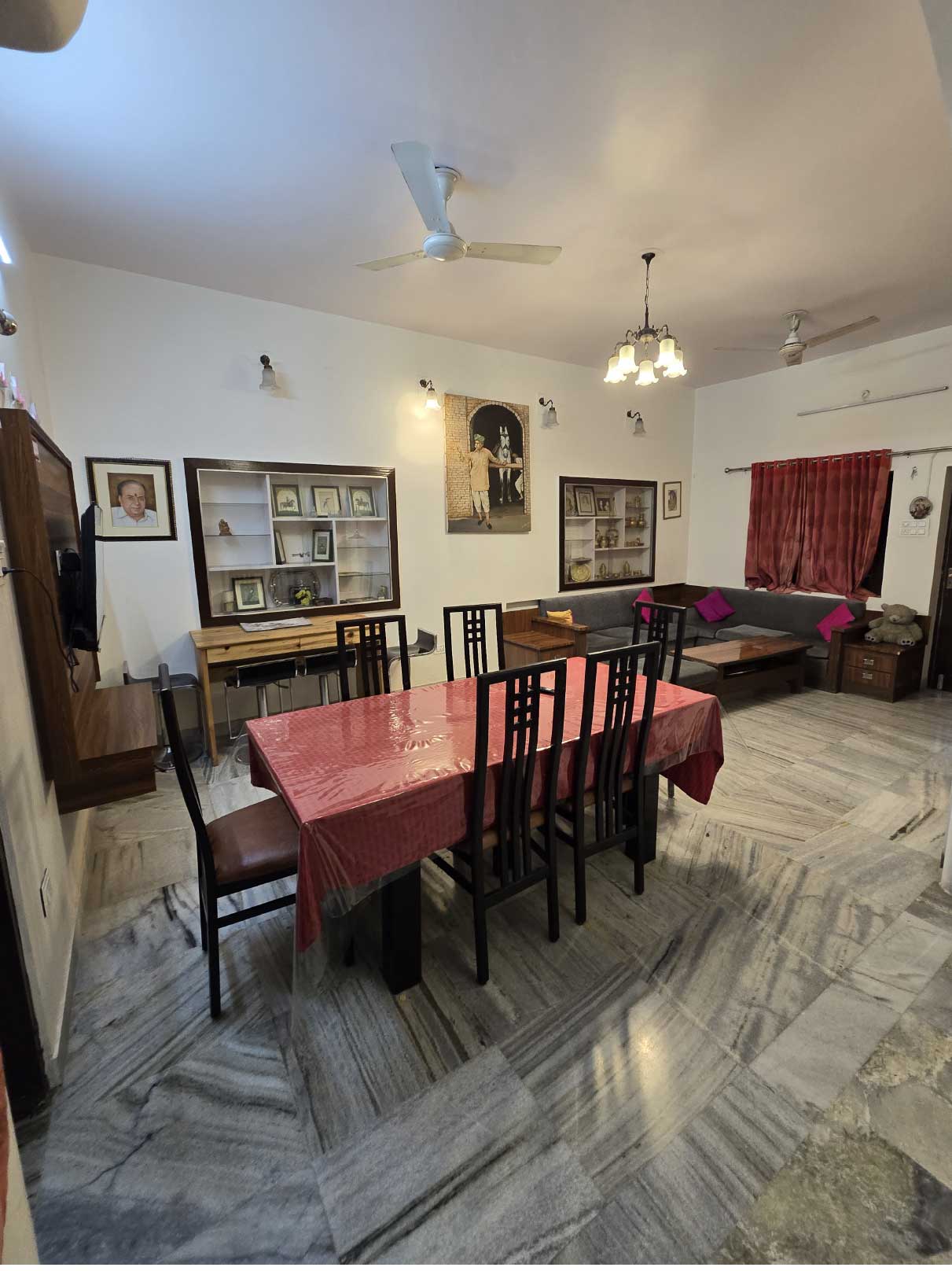 A dining area with a long dining table, several chairs, and red curtains. The room is well-lit with marble flooring.