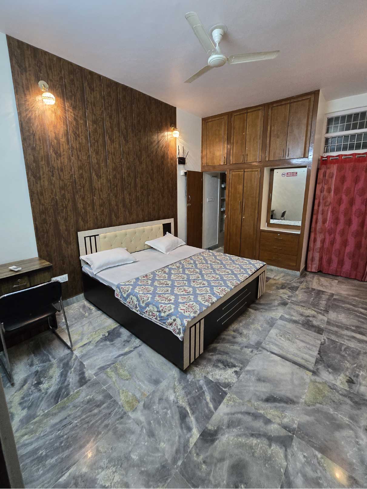 A spacious bedroom with a double bed, wooden furniture, a marble floor, and a ceiling fan. The room has bright lighting and a large wardrobe.