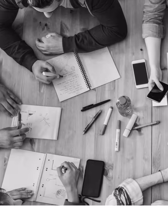 Mehrere Personen arbeiten gemeinsam an Skizzen und Notizen auf einem Holztisch, umgeben von Smartphones, Stiften und einer Wasserflasche.