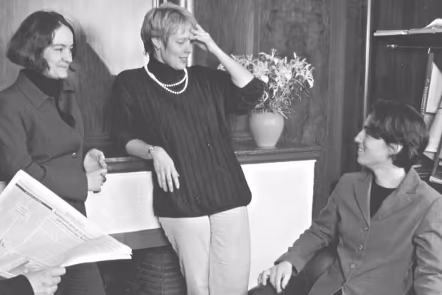 Schwarz-weiß-Foto von drei Frauen, die in einem Raum stehend und sitzend miteinander sprechen, es liegt eine Zeitung in der Hand einer Person.