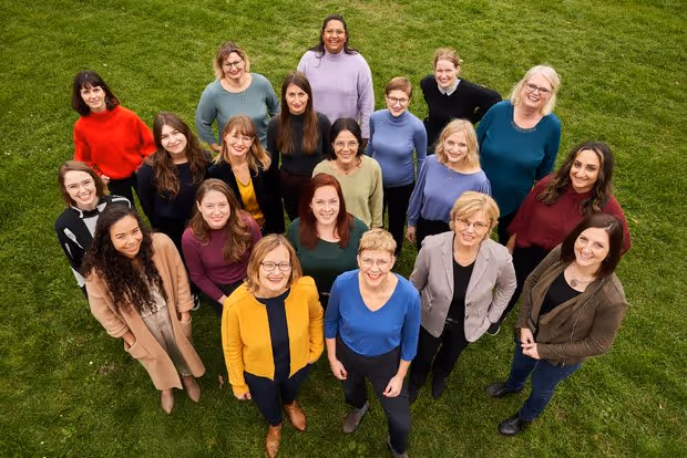 Gruppenfoto von 18 Frauen, die auf grünem Gras stehen und nach oben lächeln.