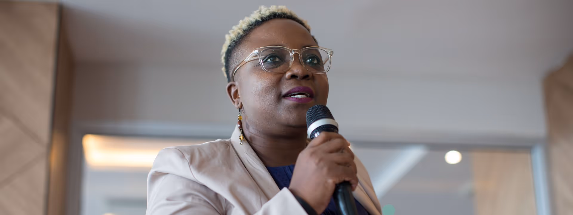 Frau mit kurzen blonden Haaren, Brille und Ohrringen spricht in ein Mikrofon.