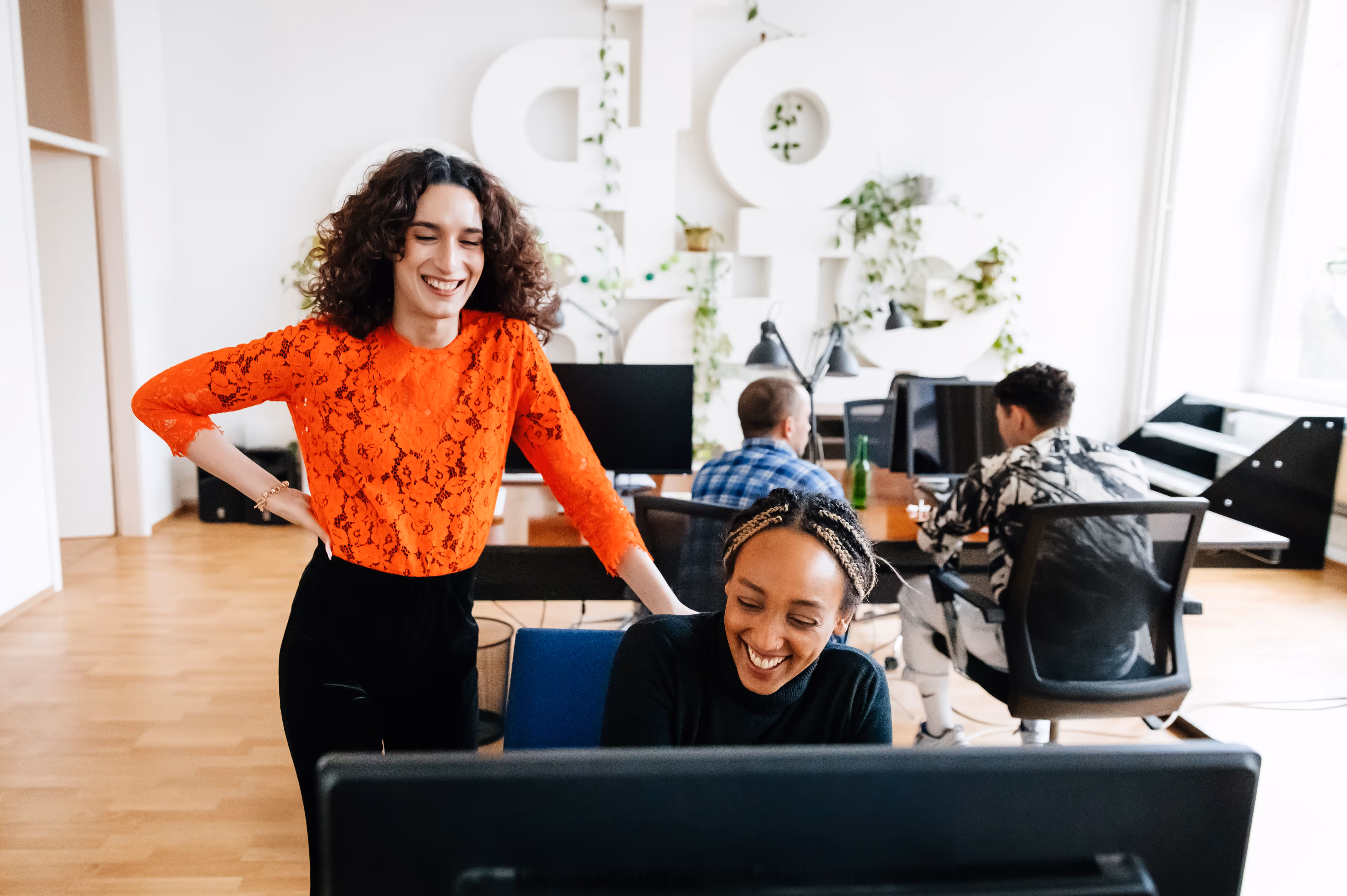 Zwei Frauen lachen zusammen vor einem Computer in einem modernen Büro, während zwei weitere Personen im Hintergrund an ihren Bildschirmen arbeiten.