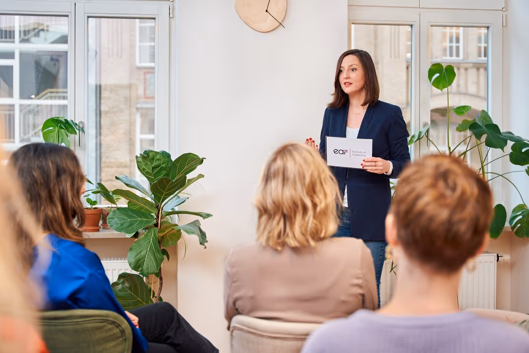 Frau hält eine Präsentation vor einem Publikum in einem hellen Raum mit grünen Pflanzen und großen Fenstern.