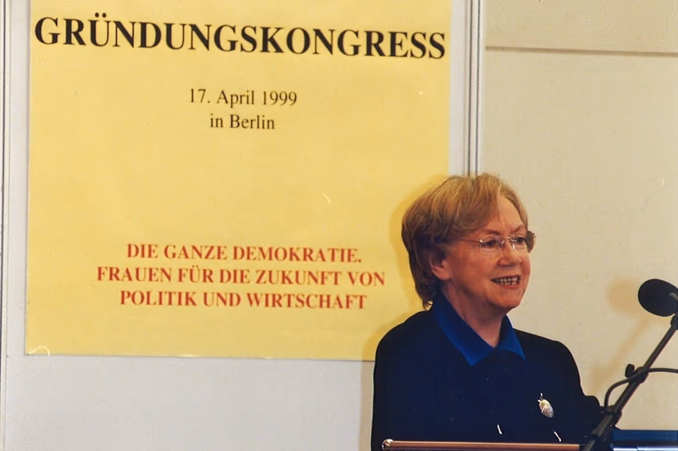 Eine Frau spricht vor einem Mikrofon neben einem gelben Banner mit der Aufschrift Gründungskongress, 17. April 1999 in Berlin, Frauen für die Zukunft von Politik und Wirtschaft.